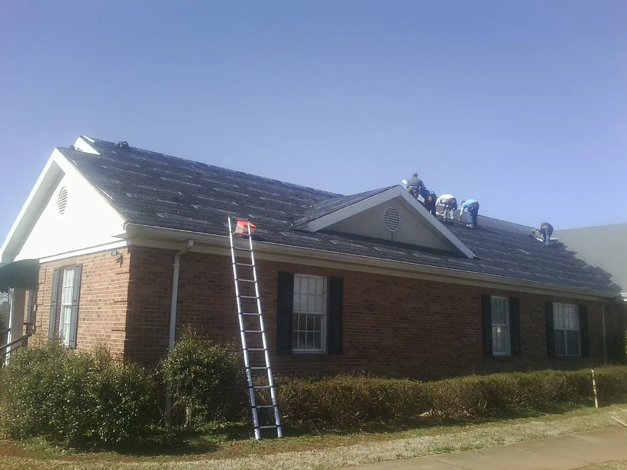 Crew tearing shingles off a brick ranch roof with a ladder set — Metal Roof Repair work by our crew in Melvin Village