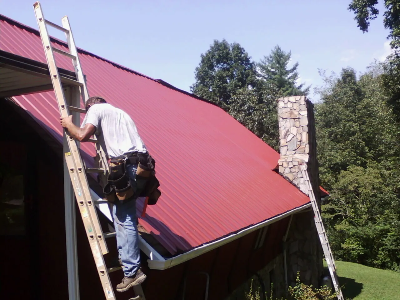 Roofer on a ladder beside a red metal roof and stone chimney — Soffit & Fascia Repair by Roofing Brothers in Melvin Village